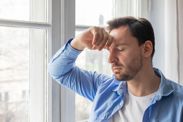 A man standing in front of a window, holding his head with his hands. His facial expression shows...