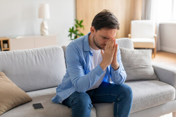 A young man is seated on a grey sofa, looking distressed with his hands covering his face. A smartphone lies nearby, suggesting he may have received upsetting news