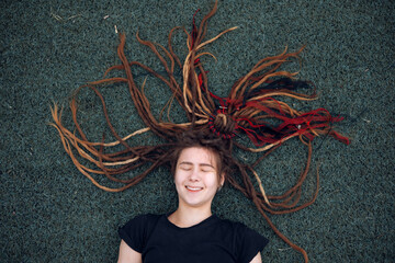 A hipster teen girl with dreadlocks lies on the grass on the playground. Daytime, stadium, open air.