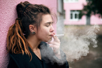 Portrait of a hipster teen girl smoking in a warm black jacket leaning on the loft brick wall,...