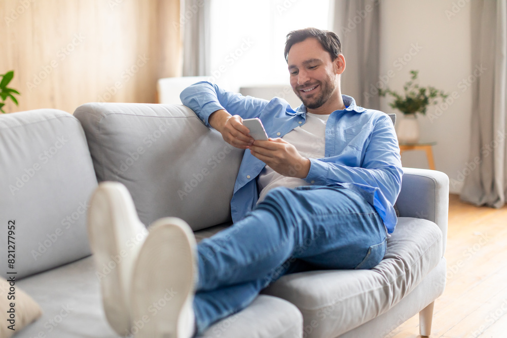 Poster happy man is reclining on a couch while actively engaged with his smartphone, focused on the screen.