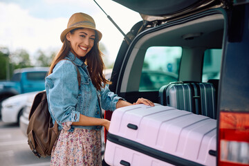 Happy traveler taking her suitcase out of car trunk on parking lot and looking at camera.