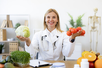 Mature female doctor nutritionist holds fresh organic tomatoes and cabbage in hands while sitting...