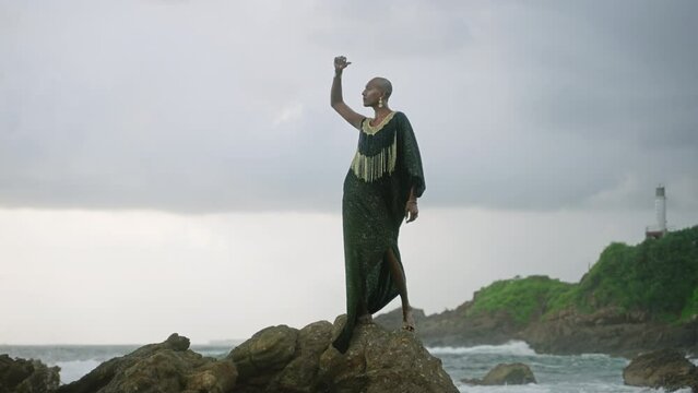 Androgynous person of color stands in boutique posh dress on rock above ocean coastline. Trans sexual ethnic fashion model in epic outfit wears golden jewellery, posing like Oshun goddess concept.