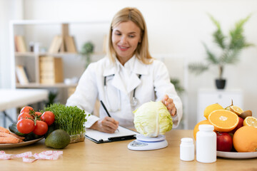 Concept of weight loss and treatment of gastrointestinal tract. Professional mature female nutritionist sitting at table in clinic and weighs cabbage, making up patient's healthy diet.