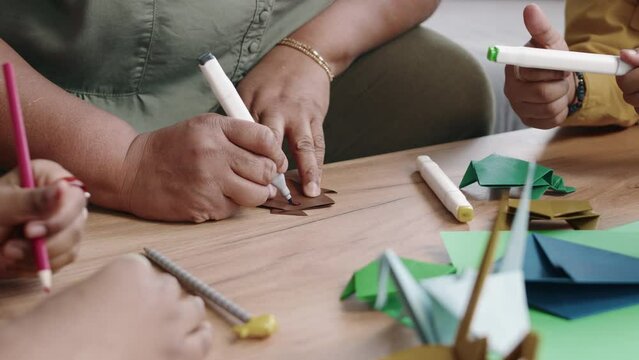 Close-up of hands of anonymous dark-skinned woman and kids carefully decorating colourful origami animals using pencils and markers, by drawing eyes