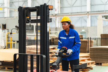 African American female worker driving forklift truck in heavy metal industrial factory, Smiling woman engineer wearing vest and helmet safety moving material at warehouse factory