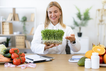 Experienced dietitian adding different vitamins, minerals and nutrients to eating. Charming Caucasian woman in doctor's coat with microgreens smiling in consulting room of private clinic.