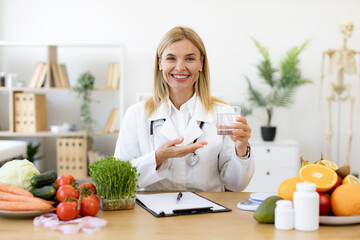 Portrait of mature lady posing with glass of clean water while sitting at writing desk in...