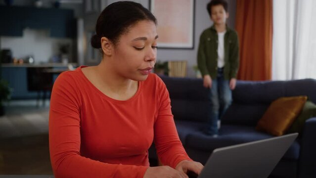 Irritated stressed African American woman freelancer mother female girl mom working distant laptop at home distracted noise hyperactive little kid boy child son jumping couch sofa say hush ask silence