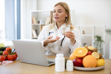 Female doctor in white coat holding glass of water during a video call. Experienced female nutritionist tells patient about benefits of water during an online session using laptop.