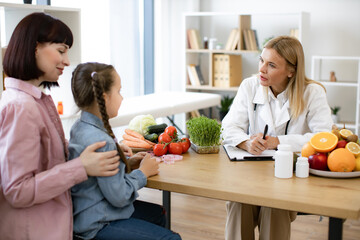 Experienced female dietitian prepares course of healthy nutrition for small child. Mature Caucasian female doctor talking with child girl sitting on her mom knees and writing diet plan.