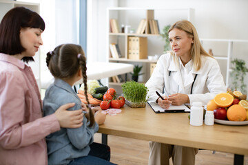 Experienced female dietitian prepares course of healthy nutrition for small child. Mature Caucasian female doctor talking with child girl sitting on her mom knees and writing diet plan.