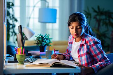 Asian Indian girl child studying at home on study table with computer, books, Globe model, victory trophy