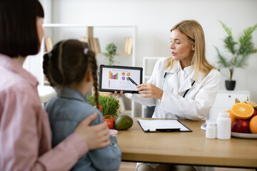 Mature Caucasian female doctor shows on tablet graphs of food for healthy eating. Little school...