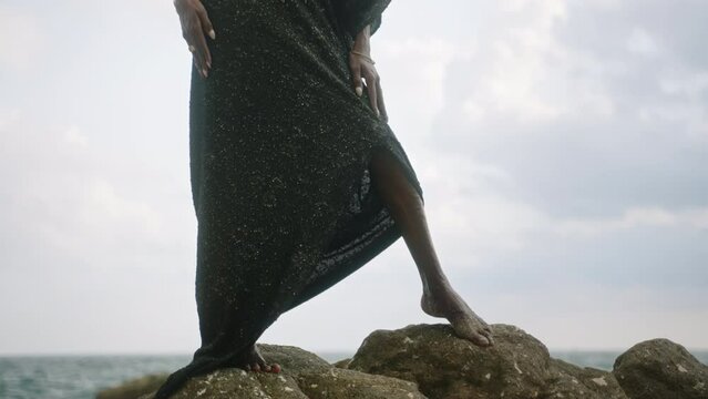 Divine non-binary black person in boutique dress, brass jewelry poses on rocky hill top above dramatic ocean cloud skyline. Queer lgbt fashion model in posh outfit fluttering in air. Pantheon concept