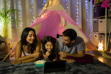 Cheerful family watching a movie at home with popcorn, surrounded by cozy lights and a fun teepee...