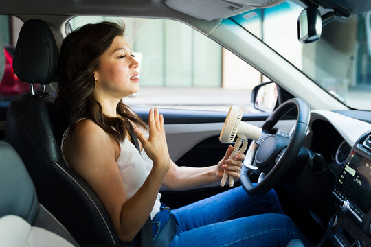 Pretty woman in her 30s holding a portable fan while sweating and driving with broken air conditioning