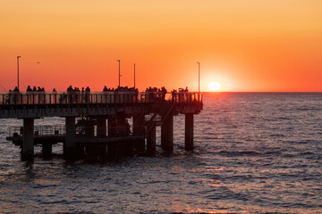 Many people watch the sunset from the pier over the sea. The orange sun was half hidden behind the...