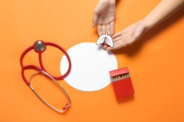 Female hands with paper lungs, cigarettes, stethoscope and blank card on orange background