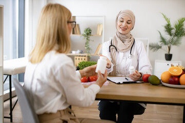 Mature Caucasian lady receives consultation on healthy eating. Muslim woman nutritionist makes plan...