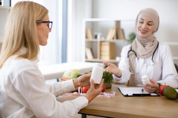 Obraz premium Mature Caucasian lady receives consultation on healthy eating. Muslim woman nutritionist makes plan for treatment of gastrointestinal tract using vitamins and supplements for healthy diet.