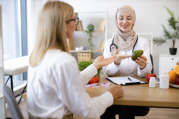 Experienced medical specialist giving advice on improving diet by abundance of nutrients. Arabian woman in hijab with fresh avocado in hand chatting with mature female lady in consulting room.