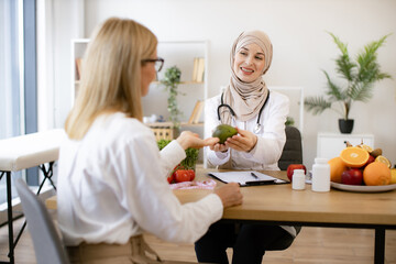 Arab female doctor makes treatment plan for gastrointestinal tract while sitting at table in modern clinic. Professional nutritionist talks to patient about benefits of healthy diet holding avocado.