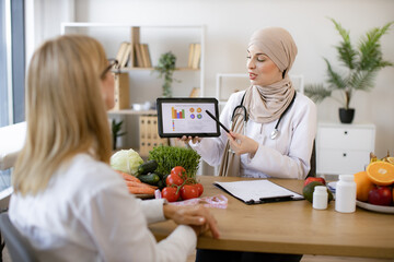 Muslim female doctor showing on tablet graphs of food for healthy eating. Mature lady patient visiting arabian woman nutritionist in hijab at modern clinic.