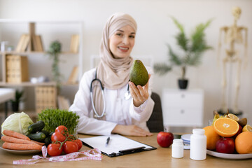 Happy female writing prescription for patients proper healthy diet. Arabian female nutritionist making healthy eating plan and calculating calorie content of avocado using weight.
