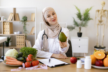 Arabian female nutritionist making healthy eating plan and calculating calorie content of avocado using weight. Happy female writing prescription for patients proper healthy diet.