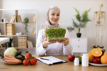 Close up view of attractive lady in white coat holding container with microgreens while sitting at desk in workplace. Muslim specialist in nutrition with hijab proposing modern addition to diet.