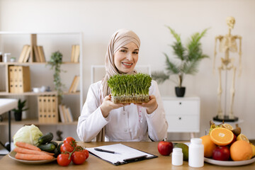 Muslim specialist in nutrition with hijab proposing modern addition to diet. Close up view of attractive lady in white coat holding container with microgreens while sitting at desk in workplace.