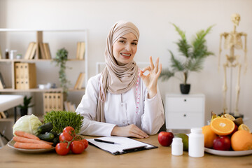 Nutritionist working writing diet plan for patient. Portrait of female nutritionist in office. Arabian nutritionist doctor with fruits and vegetables in office. Smiling young dietician sitting at desk