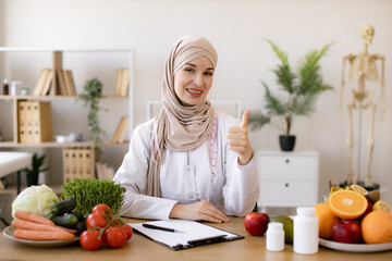 Smiling young dietician sitting at desk. Arabian nutritionist doctor with fruits and vegetables in office. Nutritionist working writing diet plan for patient. Portrait of female nutritionist in office