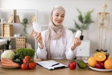 Cheerful expert in food proposing dietary supplements to improve healthy meals. Portrait of positive Muslim lady in hijab and lab coat smiling at camera while demonstrating two pills bottles in office