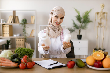 Portrait of positive Muslim lady in hijab and lab coat smiling at camera while demonstrating two pills bottles in office. Cheerful expert in food proposing dietary supplements to improve healthy meals