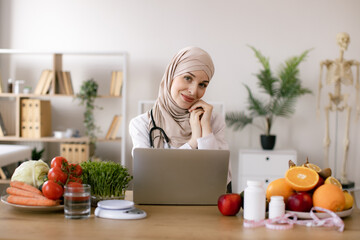 Nutritionist working on laptop and writing diet plan for patient. Muslim nutritionist doctor with fruits and vegetables in office. Smiling young dietician sitting at desk.