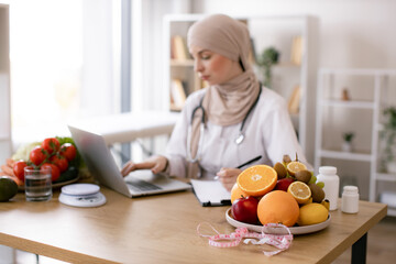 Serious Muslim lady in doctor's coat typing on modern laptop in consulting room of medical center. Experienced nutrition professional searching for weight loss information on food websites.