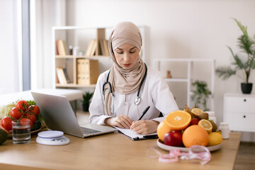 Experienced nutrition professional searching for weight loss information on food websites. Serious Muslim lady in doctor's coat typing on modern laptop in consulting room of medical center.
