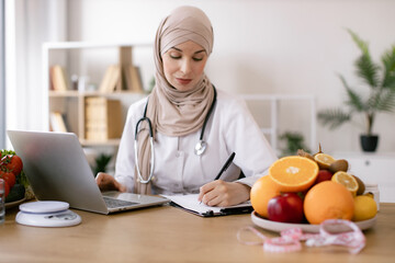 Experienced Muslim female nutritionist sitting and working with laptop in modern clinic. Young female doctor prescribes online prescription for patient healthy diet.