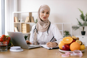 Young female doctor prescribes online prescription for patient healthy diet. Experienced Muslim female nutritionist sitting and working with laptop in modern clinic.