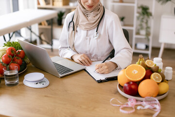 Close up of hands of Muslim female nutritionist sitting and working with laptop in modern clinic. Young female doctor prescribes online prescription for patient healthy diet.
