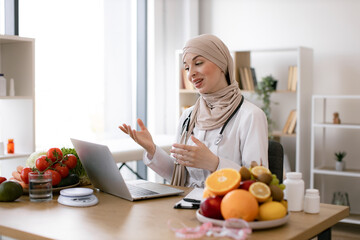 Friendly Muslim female making video call via wireless device at doctor's workplace in hospital. Attentive dietitian providing online consultation about client's progress in identifying foods.
