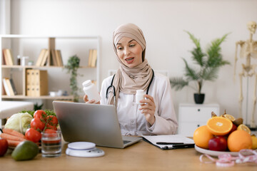 Positive arabian nutritionist woman holding pills or vitamins in hands during online consultation with patient using laptop. Muslim doctor in hijab distantly describes the treatment plan.