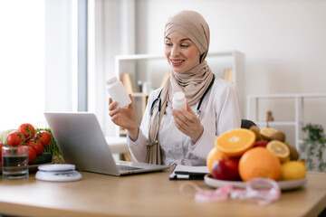 Muslim doctor in hijab distantly describes the treatment plan. Positive arabian nutritionist woman holding pills or vitamins in hands during online consultation with patient using laptop.