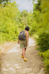 Middle aged woman dressed in shorts walking along forest path with backpack. Attractive female traveler relaxing and enjoying nature on hiking trail. Concept of mental detox and physical health