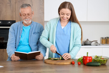 Senior man reading book with nurse cooking in kitchen