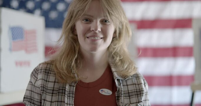 Young woman with red hair puts on "I voted" sticker and smiles. Voting booths and United States flag on back wall in background. 4K DCI