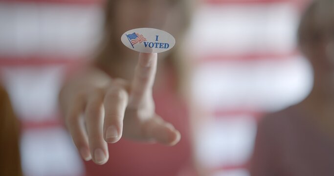 Young woman holds up oval "I voted" sticker on finger, pushed towards camera. Shallow depth of field with focus on sticker and people blurred